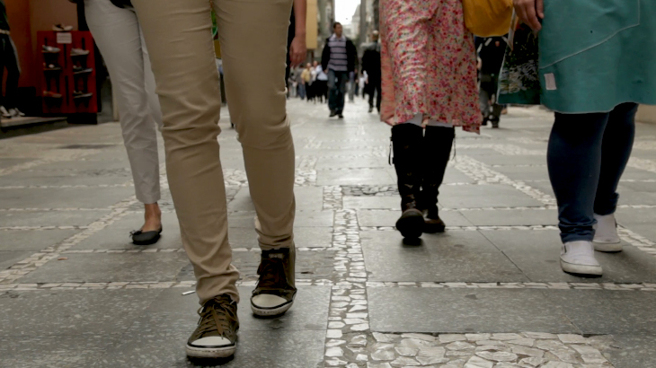 Descrição (Foto uma calçada com 4 pessoas caminhando, foto mostra apenas os membros inferiores, a partir da coxa, com o fundo desfoca)