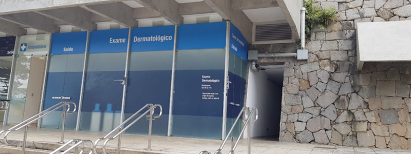 Fotografia mostra entrada da sala do exame dermatológico, com portas de vidro e adesivos azuis de identificação. Ao lado, uma parede de pedras.