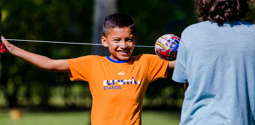 Criança com camiseta laranja do Curumim brinca com outra criança, de costas