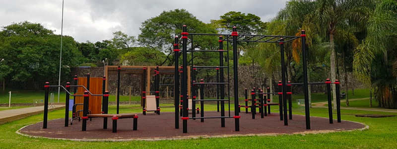 Fotografia mostra diversos equipamentos para ginástica, com barras em diferentes alturas e inclinações e tábuas de madeira. A estação fica na Arena Praia, em meio às árvores e próxima ao campo de futebol.