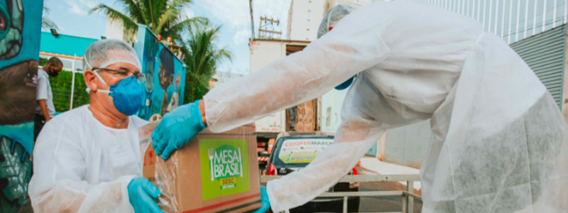 Fotografia mostra duas pessoas com avental, touca, máscara e luvas. Uma entrega para a outra uma caixa de papelão identificada com a marca Mesa Brasil.