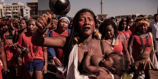 Fotografia mostra mulher indígena em primeiro plano carregando um bebê, que mama em seu peito. Ela tem um chocalho na mão e se manifesta junto de uma multidão atrás dela.