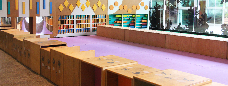 Espaço com piso de E.V.A. roxo cercado por cubos modulares de madeira com desenhos de rostos e uma parede com diversos elementos lúdicos coloridos ao fundo