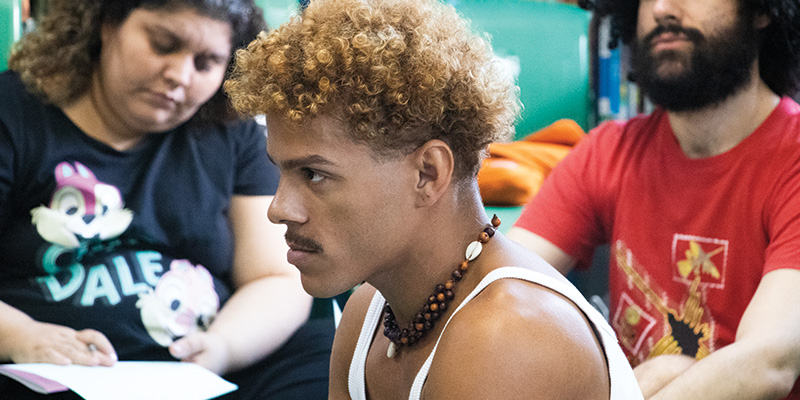 Numa roda, Franco Fonseca conversa com professores de uma escola de Ensino Fundamental. Franco é um jovem negro, com cabelos curtos e pintados de amarelo. Na foto ele aparece de perfil. Ao fundo, imagem de pessoas que acompanham o evento.