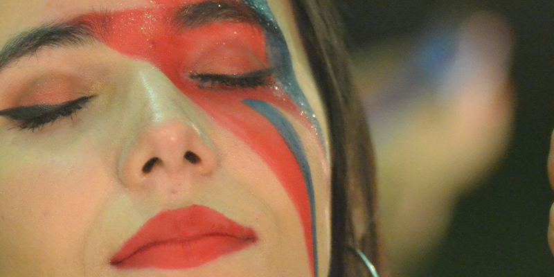 Rosto de uma mulher, de olhos fechados, maquiada com o famoso raio de David Bowie azul e vermelho.