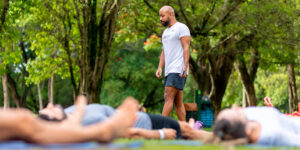 Fotografia colorida com diversas pessoas deitadas sobre a grama. Em segundo plano, uma homem negro de pele clara, o professor Pedro Costa, caminha entre as pessoas com árvores ao fundo.