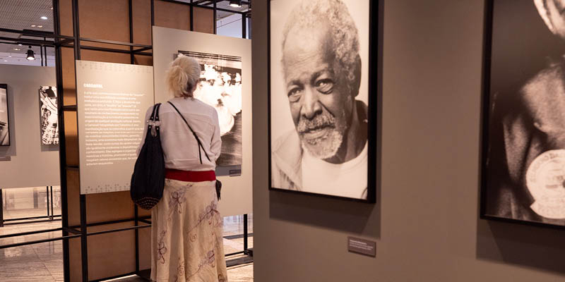Fotografia mostra uma mulher de costas visitando a exposição Consteleção Celestina. Em primeiro plano, há uma fotografia em preto e branco de um homem negro.
