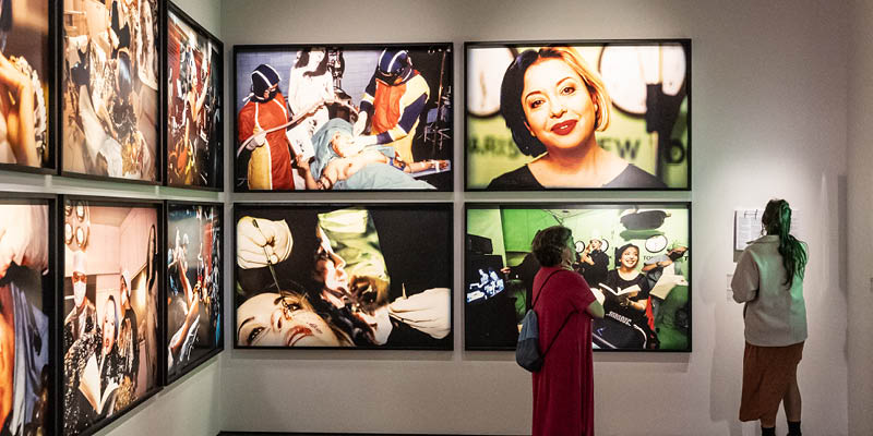 Duas mulheres observam uma galeria de fotografias em grandes proporções da artista francesa ORLAN. As fotografias mostram performances em que ORLAN passa por cirurgias plásticas.