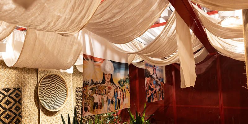 Fotografia mostra uma das salas da exposição: há diversos panos em tons crus translúcidos colocados nas vigas do teto. Nas paredes, cestarias e pinturas em tecido. No chão, plantas.