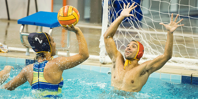 Na imagem, uma mulher usando uma toca com o número 2 e um maiô azul e amarelo prepara-se para arremessar uma bola de polo aquático em direção ao gol. À sua frente, um homem usando uma toca vermelha e amarela ergue os braços, na tentativa de depender a meta. Ambos estão dentro da piscina com água na altura do peito