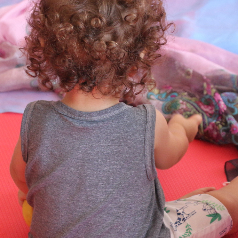 bebê de cabelos encaracolados brincando sentado, de costas para a câmera.