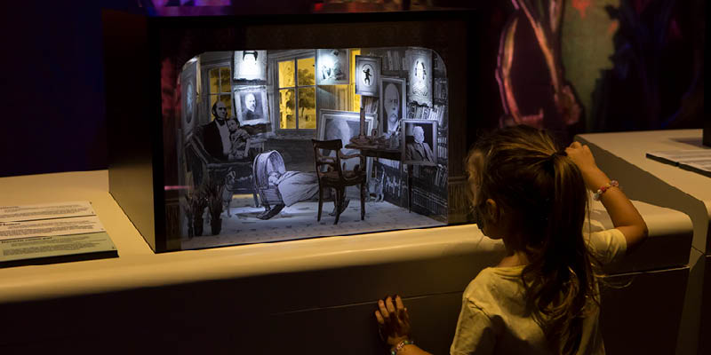 Fotografia mostra uma menininha observando a maquete iluminada do escritório do cientista Charles Darwin.