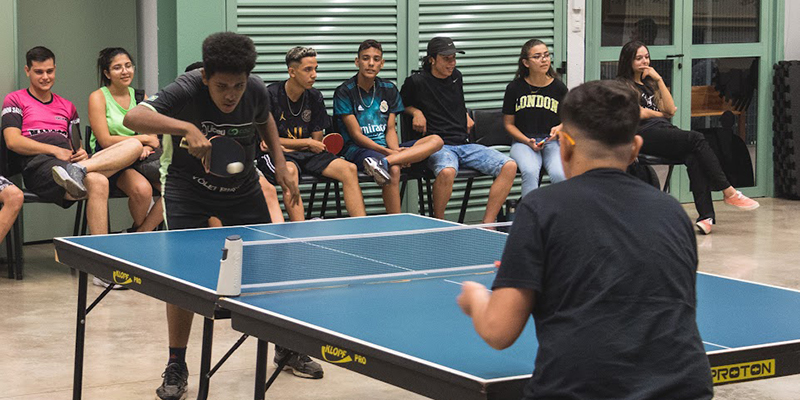 Dois jovens estão jogando tênis de mesa em uma sala com várias pessoas sentadas ao fundo observando.