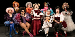 A imagem mostra um grupo de dez artistas performáticos, todos vestidos com figurinos elaborados e maquiagens chamativas. Cada um dos artistas tem uma expressão facial distinta e está usando roupas com muitos detalhes como rendas, perucas volumosas, joias e botas altas, criando uma cena visualmente impactante e cheia de personalidade.