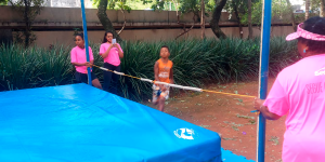 A imagem mostra uma criança correndo em direção a uma área com um colchão azul, delimitada por duas hastes verticais com uma faixa elástica estendida entre elas. Três mulheres vestidas com camisetas rosa estão próximas: uma segura a faixa elástica de um lado, outra do lado oposto e a terceira, ao fundo, segura um celular apontado para a criança. Ao redor há vegetação e um chão de terra batida.