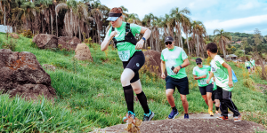 A imagem mostra pessoas correndo em trilha com vegetação e pedras. Em destaque, uma mulher com viseira, camiseta verde (número 112), bermuda preta e meias até o joelho sobe uma pedra. Atrás dela, outros participantes também usam camiseta verde e seguem o percurso. Ao fundo, há palmeiras e mais corredores.