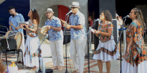 Seis pessoas estão posicionadas em uma linha, em pé, segurando instrumentos musicais e microfones. Três delas estão com microfones próximos à boca. Os instrumentos visíveis incluem tambor, ganzá e pandeiro. Duas pessoas usam chapéus. As roupas do grupo têm estampas florais e cores similares. No chão, à frente do grupo, há um banco e uma cabaça. Ao fundo, há uma parede cinza com um arco e outras pessoas parcialmente visíveis.