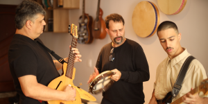 Três músicos estão em um ambiente fechado, ensaiando juntos. À esquerda, um homem de camiseta preta toca uma guitarra elétrica de madeira clara com formato estilizado. Ao centro, outro homem de roupa preta toca um pandeiro e parece estar cantando ou falando, com expressão concentrada. À direita, um jovem de camisa clara toca um baixo elétrico. No fundo, vários instrumentos musicais estão pendurados na parede — entre eles, violões e pandeiros — e um amplificador Marshall está posicionado no chão.