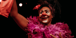 Artista caracterizada como palhaça está em cena, usando nariz vermelho, maquiagem cômica, adereço de plumas rosa e faixa com penas na cabeça. Ela levanta um pano vermelho com uma das mãos. O fundo é escuro e a iluminação destaca o rosto.