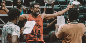 Homem de óculos e barba grisalha está de braços abertos, vestindo uma regata vermelha com estampa e texto. Ele está em ambiente interno com arquibancadas ao fundo, falando com duas pessoas que seguram folhas de papel. Ao fundo, há mais pessoas e instrumentos de percussão visíveis.