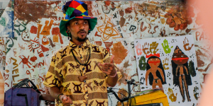 Pessoa usa chapéu colorido e camisa estampada enquanto gesticula com as mãos em frente a uma parede coberta por pinturas infantis. Atrás dela, há cartazes com desenhos feitos com tinta, incluindo figuras humanas e marcas de mãos. No canto inferior esquerdo, há caixas coloridas empilhadas e um microfone.