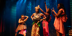 Quatro artistas estão no palco, em poses expressivas e coreografadas, usando figurinos coloridos com estampas africanas e tênis. A iluminação quente e o fundo com padrões geométricos ressaltam a estética afro-brasileira da apresentação.