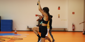 A imagem mostra um grupo de pessoas praticando atividade física em ambiente fechado com piso laranja. Em primeiro plano, uma mulher de cabelo preso, vestindo top e shorts esportivos pretos, levanta os braços com um pequeno peso em cada mão enquanto ergue um joelho. Ao fundo, outras pessoas também estão envolvidas na atividade, realizando movimentos semelhantes. No canto esquerdo inferior há vários bambolês coloridos no chão e um cone de sinalização. Na parede do fundo há um extintor de incêndio, placas de sinalização e colchonetes empilhados.