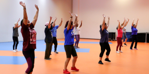 Grupo de pessoas, majoritariamente mulheres idosas, pratica atividade física em ambiente interno com piso laranja e azul. Todas estão com os braços erguidos, em posição de exercício coletivo, distribuídas em fileiras. Elas vestem roupas esportivas variadas e aparentam estar acompanhando uma rotina de movimentos coordenados.
