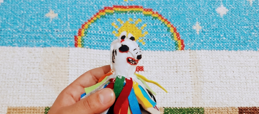 A imagem exibe um boneco artesanal da La Ursa colorido sobre um bordado de arco-íris e sol
