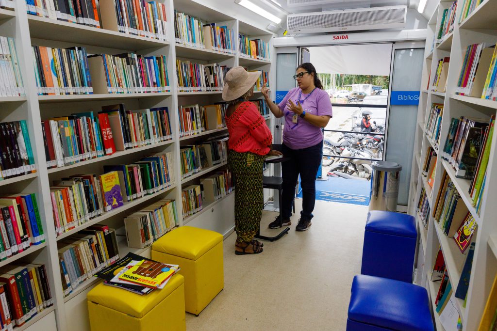 Fotografia mostra duas mulheres conversando dentro de uma carreta do BiblioSesc. Há pufes no chão e as paredes são tomadas por prateleiras com livros. Há iluminação e climatização.
