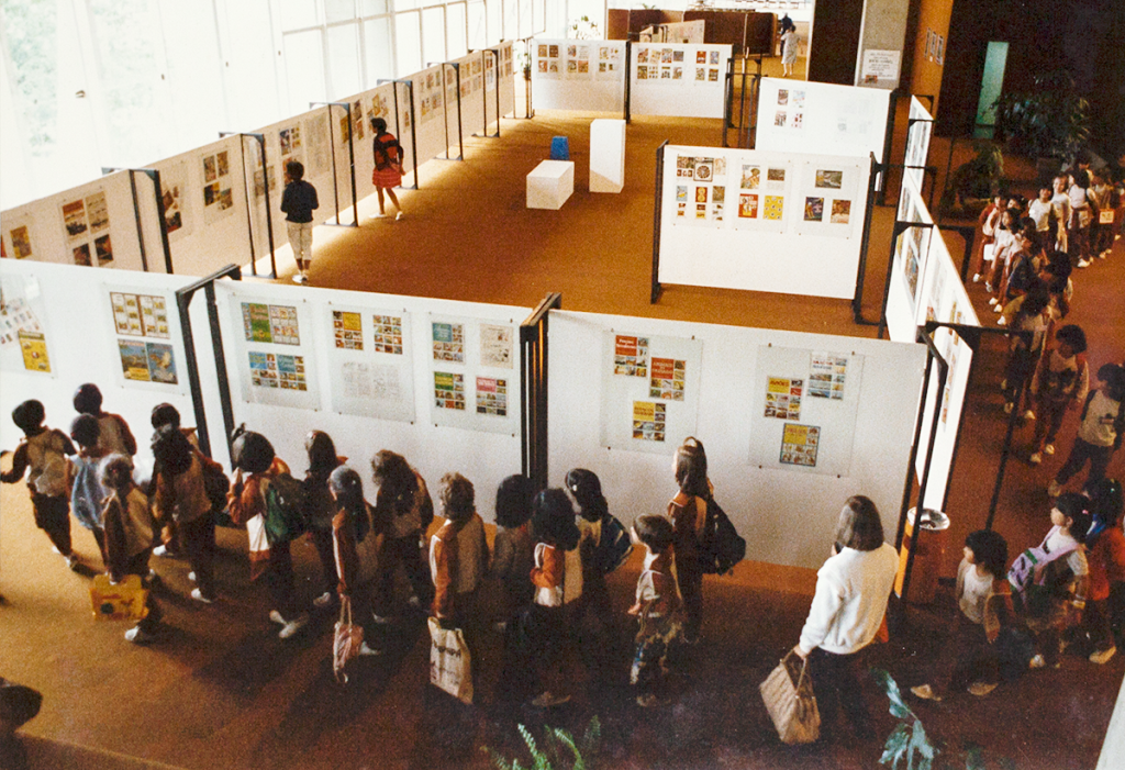 Foto com vista superior de espaço expositivo com chapas brancas onde estão afixadas reproduções de álbuns de figurinhas. Uma fila ordenada de crianças percorre a exposição.