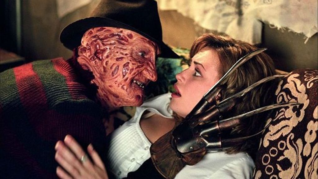 A imagem mostra uma cena tensa e intimidadora entre dois personagens. À esquerda, está um homem com o rosto gravemente desfigurado, coberto por queimaduras profundas, cicatrizes e pele avermelhada e irregular. Ele usa um chapéu preto de aba larga e um suéter de tricô com listras vermelhas e verde-escuras. Seu olhar é fixo e ameaçador, e ele está inclinado sobre uma jovem. À direita, uma mulher jovem de cabelos castanhos claros está deitada de costas, olhando para o homem com uma expressão de medo e surpresa. Ela veste uma blusa branca de botões. O homem está usando uma luva metálica com longas lâminas afiadas, que estão posicionadas muito perto do rosto e do pescoço da mulher, criando uma sensação de perigo iminente. A mão esquerda do homem, sem a luva, aparece pressionando o ombro da mulher. O fundo é escuro e um pouco desfocado, sugerindo um ambiente interno, talvez um quarto ou sala, com uma textura que lembra um estofado ou parede. A iluminação é dramática, destacando os rostos dos dois personagens e a lâmina da luva. A cena transmite uma forte sensação de suspense e horror.